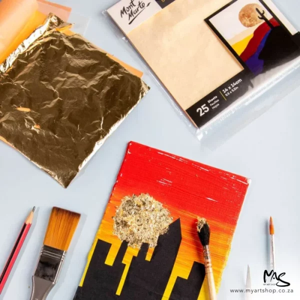 A promotional image for Mont Marte Imitation Gold Leaf. There is a sealed pad and an open pad in the top of the frame with a picture of a building in a sunset at the bottom of the frame and the gold leaf is being applied to the picture using a paint brush.