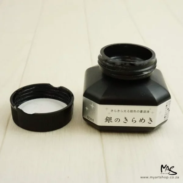 An open bottle of Silver Kuretake Mica Ink can be seen in the center of the frame horizontally. The lid is to the left of the bottle. It is on a light wood surface.