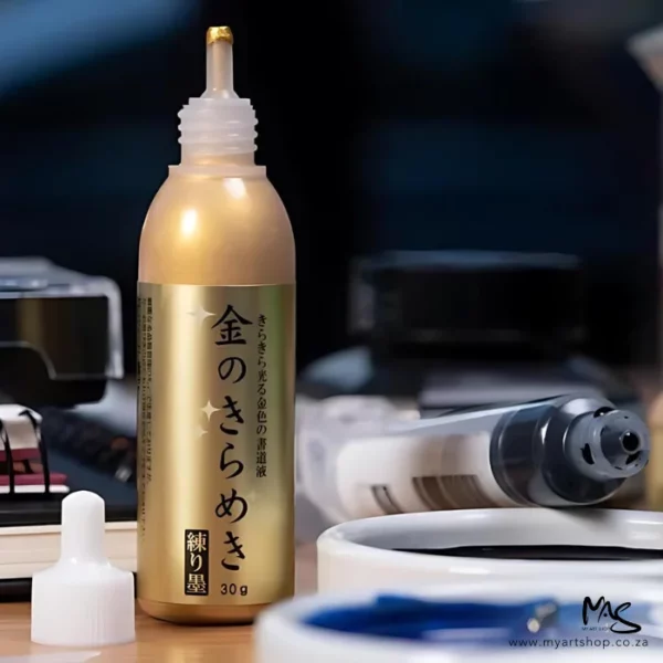 A close up of a bottle of Gold Kuretake Mica Paste can be seen standing on a desk. The lid is off and set to the left of the bottle. The bottle is aligned more to the left hand side of the frame. The bottle is clear plastic with a gold label that has black chinese print on the label. There are other art supplies in the background.