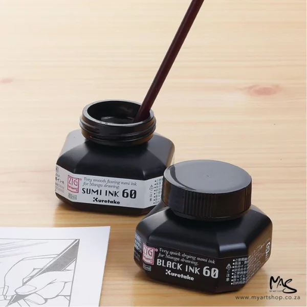 There are two bottles of ink sitting on a wooden table. One bottle is ZIG Cartoonist Sumi Black Ink and does not have a lid on it. There is a brush sitting in the ink. The other bottle is sealed. Part of a picture can be seen in the bottom left hand corner of the frame which has been drawn, using the ink.