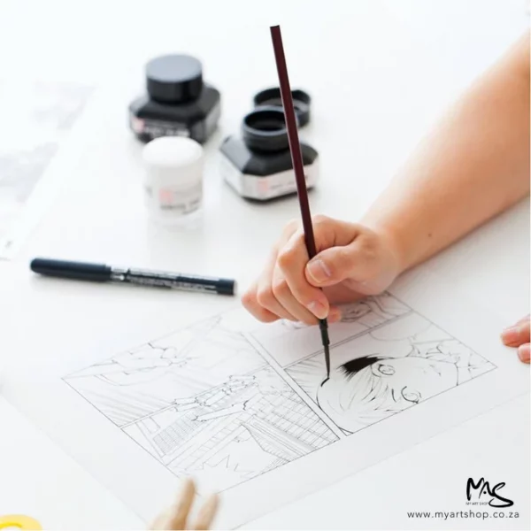 A person is drawing using ZIG Cartoonist Sumi Black Ink. They are using a paint brush to draw comics. There are bottles of ink in the background which are blurred.