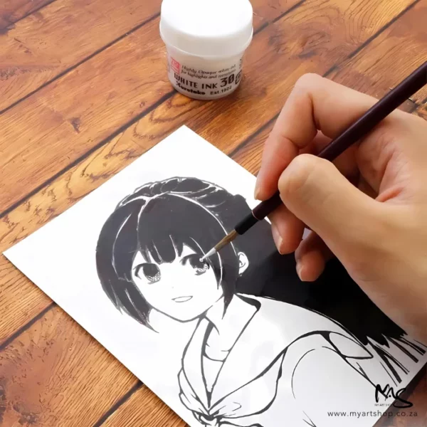 A close up of a persons hand drawing a cartoon using ZIG Cartoonist White Ink. They are using the white ink on a paint brush to add details to the eyes. There is a bottle of the ink in the background. They are working on a wooden desk.