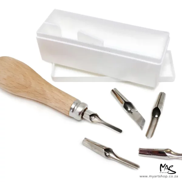 An open set of ABIG Lino Cutting Tools can be seen in the frame. The white plastic box is in the background, which houses the handle and blades. The wooden hand is to the left of the frame with one metal blade attached and the remaining four metal blades are scattered around the handle. On a white background.