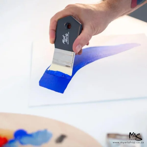 A close up of a persons hand using the Mont Marte Abstract Expression Brush 50mm. Their hand is coming out of the right hand side of the frame and they are painting a blue line using the brush. The brush has a grey handle with the Mont Marte logo printed on it.