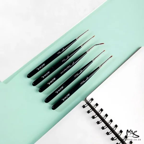 The brushes from the Mont Marte Mini Detail Brush Set 5 Piece are shown loose in the frame. They are laying diagonally across a teal strip and there is a canvas above them and an open spiral bound notebook below them.