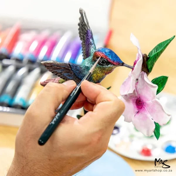 A promotional image for the Mont Marte Mini Detail Brush Set 5 Piece. A persons hand is seen coming out of the bottom of the frame holding a fine detail brush, they are painting a hummingbird ornament using the brush. There is a blurred image of a paint set in the background.