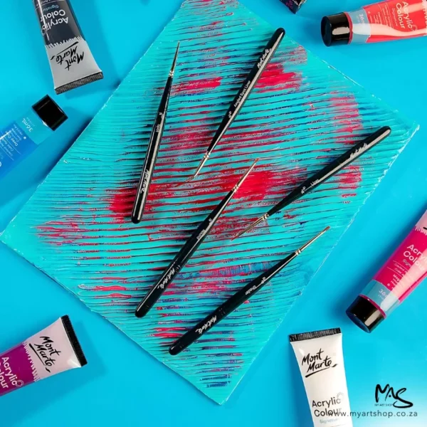 A promotional image for the Mont Marte Mini Detail Brush Set 5 Piece. The 5 brushes are laying on top of a gelli plate and there is paint randomly on the plate. The image is on a blue background and there are various tubes of Mont Marte paint scattered around the plate.