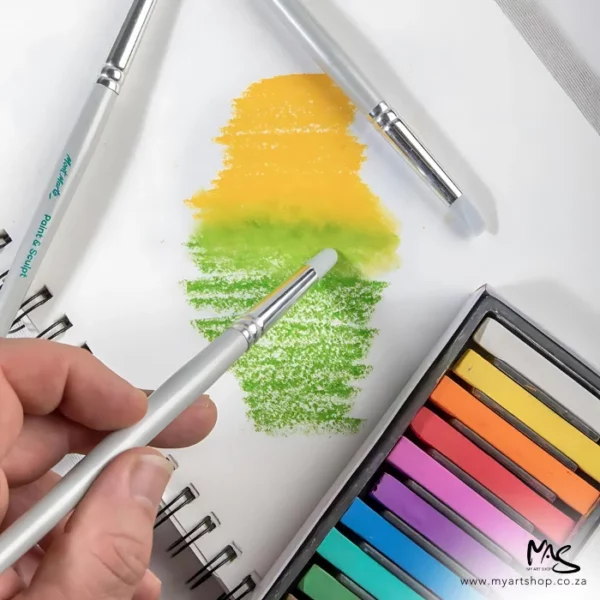 A single paint shaper from the Mont Marte Paint and Sculpt Shaper Set 5 piece is seen in a persons hand, being used to manipulate chalk on a piece of paper - to blend it. The chalk is green and yellow. You can see part of a set of coloured chalks in the bottom right hand corner of the frame.