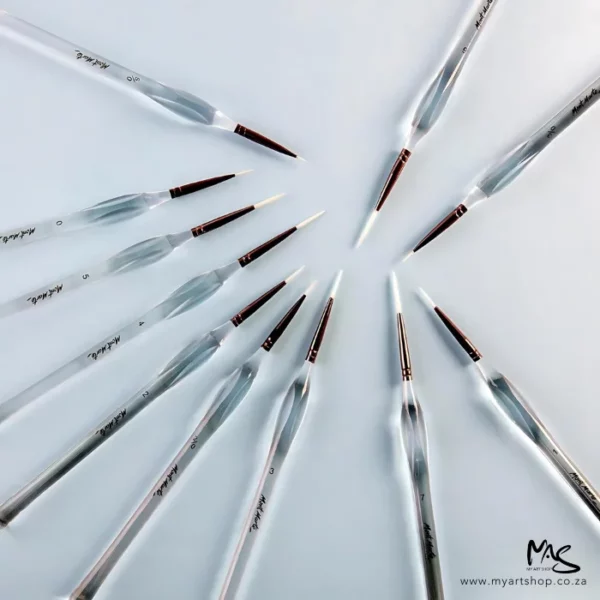 The loose brushes from the Mont Marte Signature Detail Brush Set 11 piece can be seen scattered around the frame randomly on a light grey background. the brushes have clear plastic barrels with the Mont Marte logo printed on them and the taklon brush bristles are white.