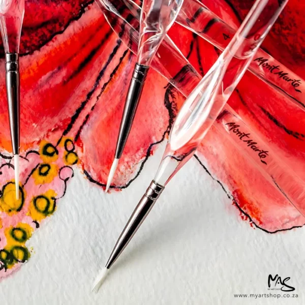 A close up of 3 brushes from the Mont Marte Signature Detail Brush Set 11 piece. The brushes have clear plastic barrels with a metal ferrule and white taklon brustles. They are laying on top of a red and yellow picture. The image is cut off by the frame.