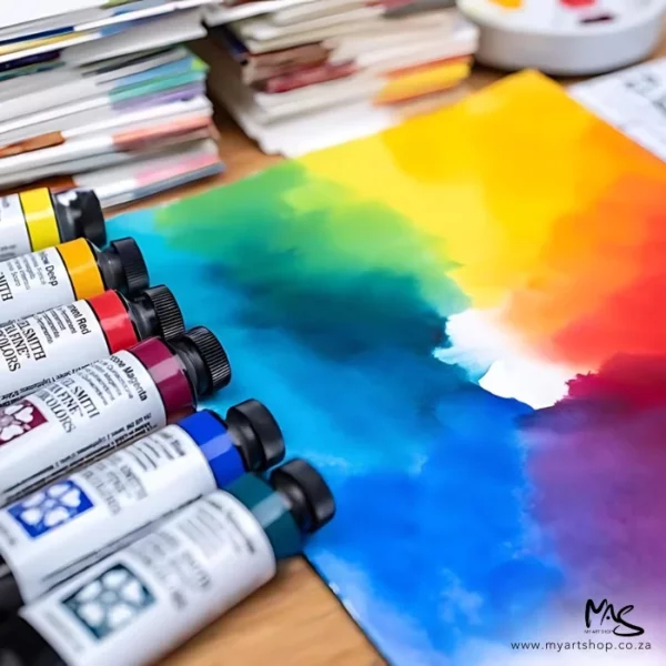 A promotional image for Daniel Smith Watercolours. There are 6 tubes coming out of the left hand side of the frame and they are laying on a piece of paper that has been painted with a rainbow of colours that are graded into each other. There are some papers in the background that are blurred.