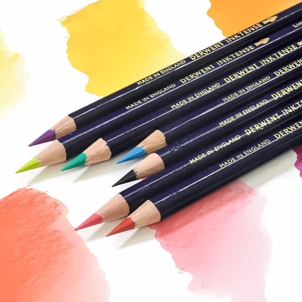 There are 7 Derwent Inktense Colour Pencils laying diagonally across the frame on a piece of paper that has various different colour swatches on it. The pencils are coming into the frame in the top right hand corner of the frame and are cut off by the frame. The pencils have a blue barrel and different coloured leads.