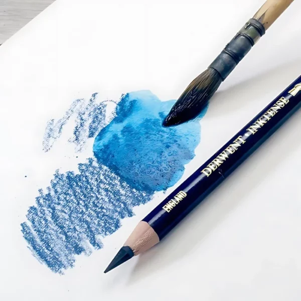 There is a single blue Derwent Inktense Colour Pencil shown laying on a piece of white paper with a sketch swatch on the paper. There is a paint brush that is being run through the swatch to show that the pencil is water soluble. A close up and is cut off by the frame.