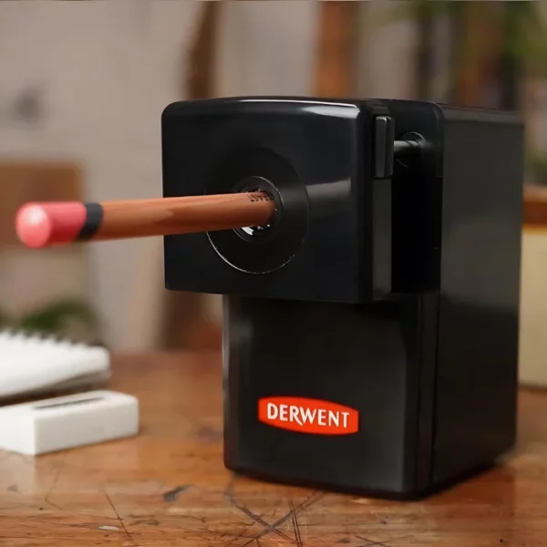 A single Derwent Super Point Mini Manual Sharpener is shown filling the center of the frame. The sharpener is black and has a handle at the back t hat turns the pencil. The red Derwent logo is printed on the pull out drawer of the sharpener. The sharpener is sitting on a wooden surface and there is a pencil in the sharpener.