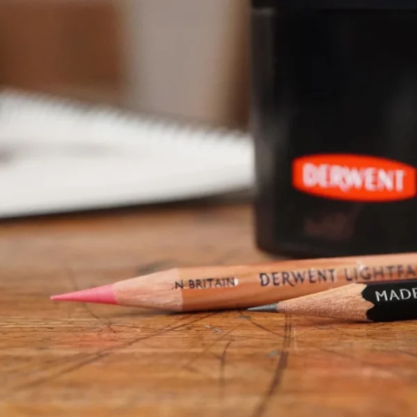 A close up of part of a Derwent Super Point Mini Manual Sharpener and two pencils that are laying next to the sharpener. The pencils have been sharpened using the sharpener and have long thin leads.