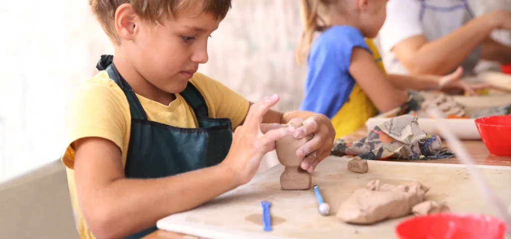 a little boy is in the left hand side of the image creating an air dry clay dinosaur out of terracotta clay. he is wearing a yellow shirt and a dark apron over it. he is molding the clay using his fingers. in the background to the right is a girl also working on a clay project. infront of him are various tools that he is using to sculpt his clay. the background is a blurred visual of the room