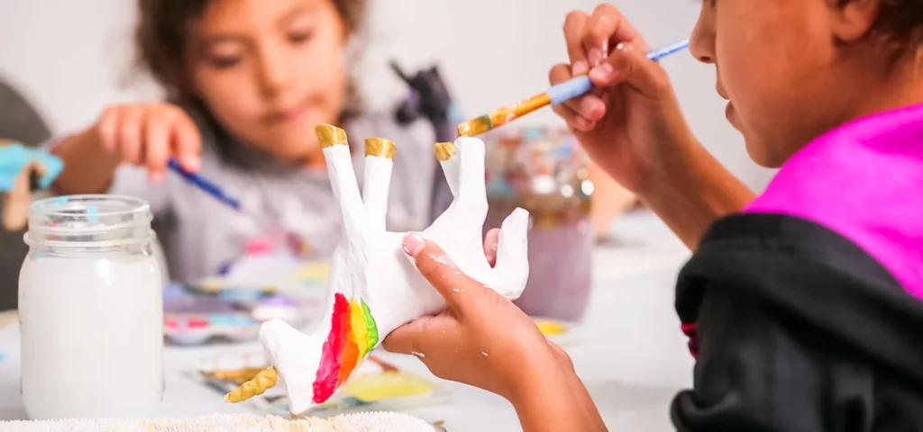 a little girl is busy painting her air dry clay unicorn. at the front of the image. she has painted its main a rainbow colour and its hooves a gold. in the background you can see the blurred face of another little girl painting her unicorn a colour, there are various items on the table but they are all very blurred. the image is very bright and happy feeling.