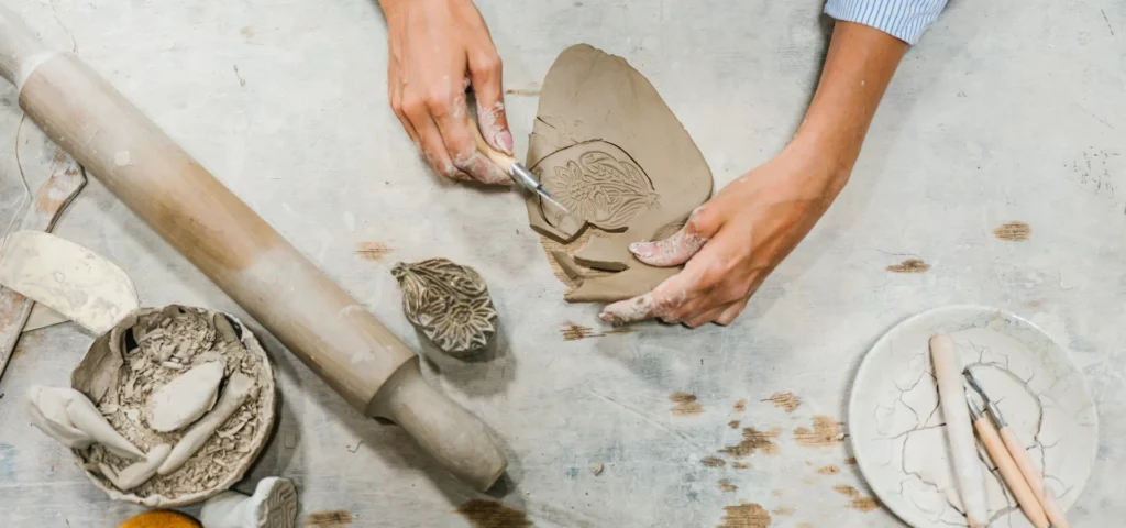 a lady is sculpting an air dry clay bowl in the center of the image. the bowl is a terracotta colour and an oval shape. she is carving patterns into it with a carving tool. on the left hand side is a rolling pin and a sponge. she is working on a table that is covered in dry clay and tools.