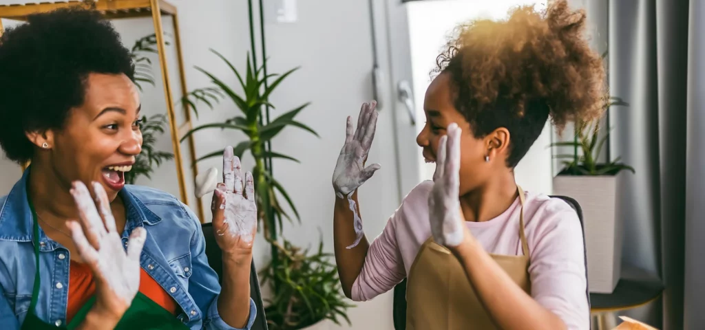 two woman are looking at eachother and are about to high five with both hands. both of their hands are covered in air dry clay. they are wearing theor normal clothes with brown aprons over them. sitting in a room with a green leafy plant in the background inbetween them.