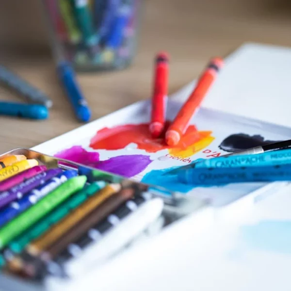 An open tin of Caran D'Ache Neocolor II Watersoluble Wax Oil Pastels with some pastels in the open lid, are shown in the frame. The pastels in the lid have been mixed with water as they are watersoluble. The background is blurred.