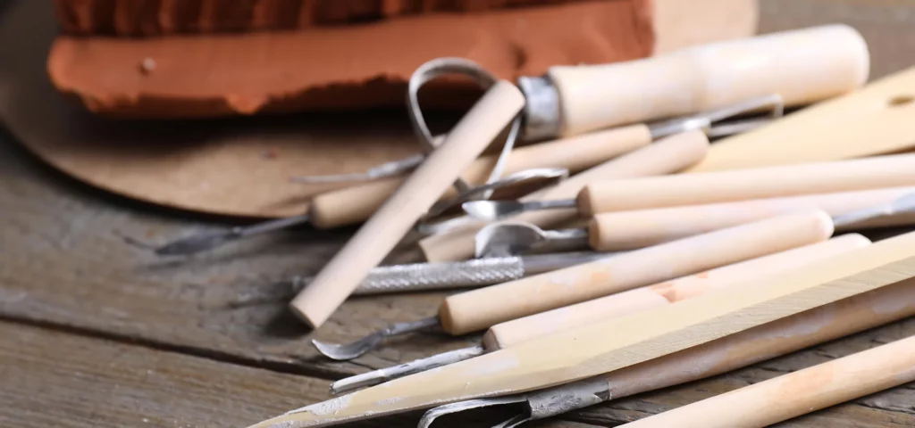 these wood and metal clay tools are laying on the side of the image facing into the left hand side. they are made of light wood and metal. there are many different types lyiong in the image and they have been used. they are all layed ontop of a table with clay that has been used int he background.