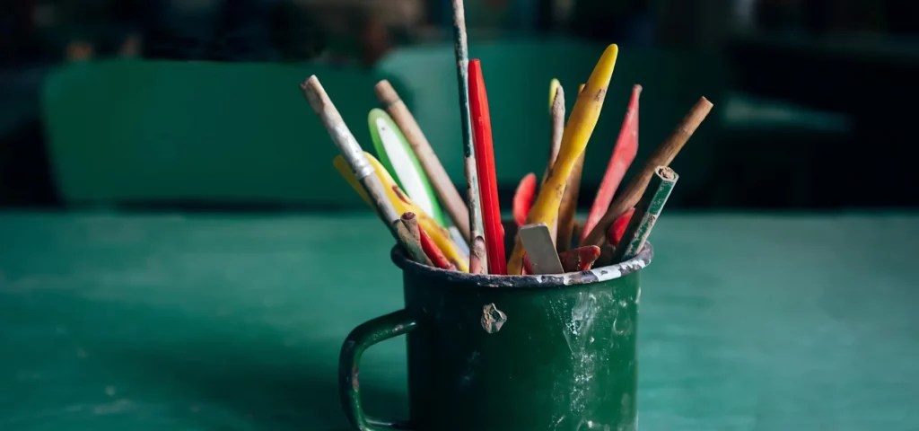 these clay tools are made of plastic and rubber and are sitting in a green mug. they are a bunch of different colours and shapes and sizes and are in the center of the image. they are right at the front of the image and the background is a blurred image of the room behind them