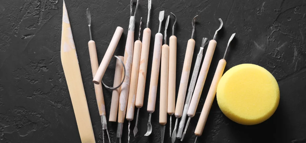 these clay tools have been scattered on the table. they all have wooden handles and are double ended. they have metal ends and are all different shapes and sizes of tools used for clay. hooks, and paddles and loops. they are lying on a flat wooden surface in the center of the image.