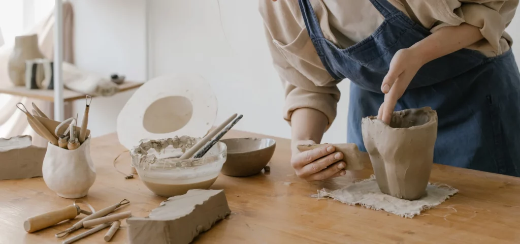 these clay tools are the smoothers. the image shows a lady on the right hand side sanding and smoothing her clay piece using a sand paper block. she is wearing a beige shirt with a blue apron and is working bent over a table with all her clay tools and clay ionfront of her. the background is a white wall