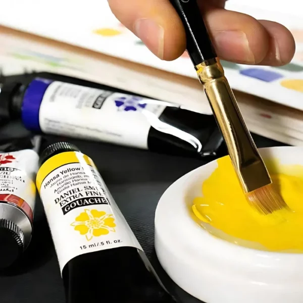 A small white ceramic bowl can be seen coming into the frame, in the bottom right hand side of the frame. There is yellow Daniel Smith Extra Fine Gouache Paint in the bowl and a persons hand is seen dipping a paint brush into the paint. There are three tubes of Gouache Paint laying around the bowl and some paper in the background. It is a close up image and the image is cut off by the frame.