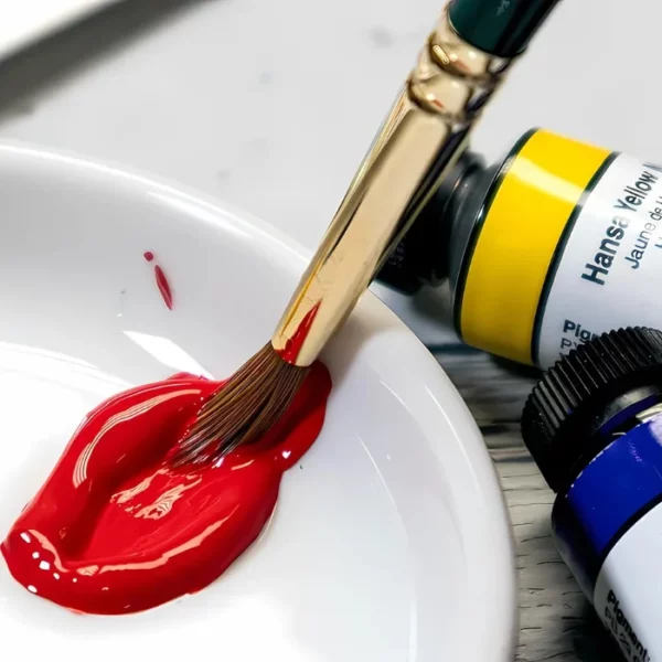 A close up of a white porcelain bowl with red Daniel Smith Extra Fine Gouache Paint in the bowl. A paint brush is seen being dipped into the paint. Part of two tubes of Daniel Smith Extra Fine Gouache Paint can be seen next to the bowl, along the right hand side of the frame. The image is cut off by the frame.