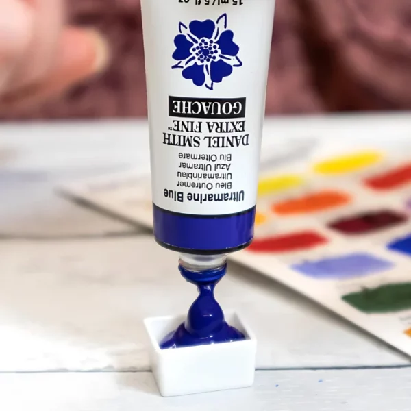 A close up of a tube of Daniel Smith Extra Fine Gouache Paint. It is shown vertically in the center of the frame, upside down, and the blue paint is being squeezed into a small white porcelain pan. The background is blurred and there is a colour swatch to the right of the frame, behind the tube.