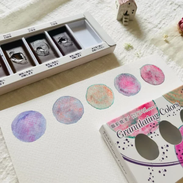 A promotional image for Granulating Gansai Tambi Watercolour Sets. You can see the open set in the background showing the paint pans in the base of the set. The box cover is shown to the front of the frame and it is lying on a piece of watercolour paper that has painted circles that were painted using the paints in the set. You can see how the colours granulate on the paper. A close up image.