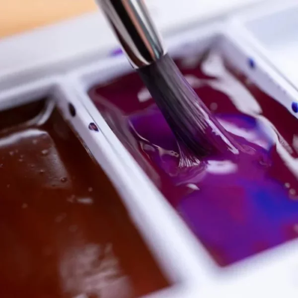 A close up of two paint pans from the Granulating Gansai Tambi Watercolour Set. A paint brush is being dipped into one of the pans wetting the colour. The pans are purple and a deep red.