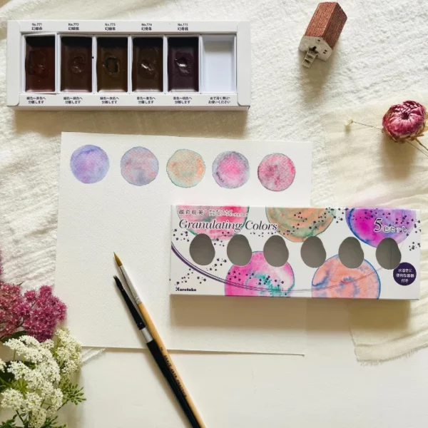 A promotional image for Granulating Gansai Tambi Watercolour Sets. You can see the open set in the background showing the paint pans in the base of the set. The box cover is shown to the front of the frame and it is lying on a piece of watercolour paper that has painted circles that were painted using the paints in the set. You can see how the colours granulate on the paper.