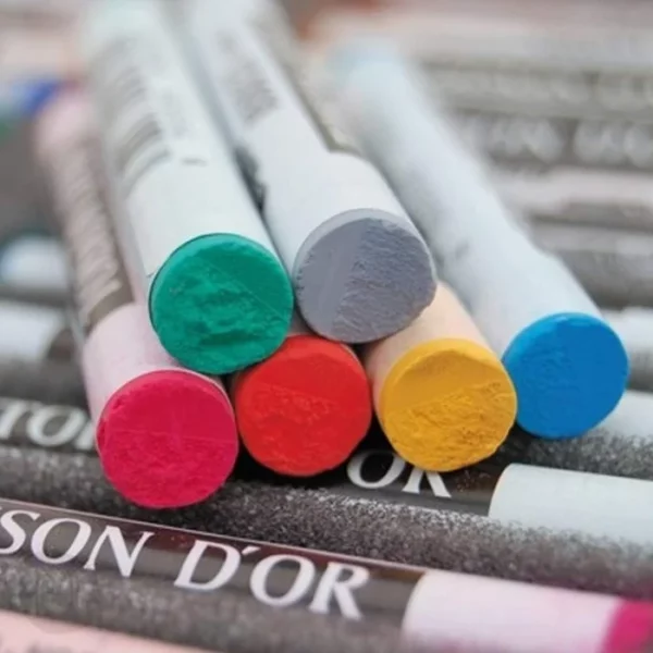 The frame is filled with a close up of various different coloured Koh-I-Noor Toison D’or Soft Pastels. There is a layer of pastels along the bottom and then a pile of pastels on top of them. They are round pastel and have a paper label around the body of the pastel with the brand name and code colour.