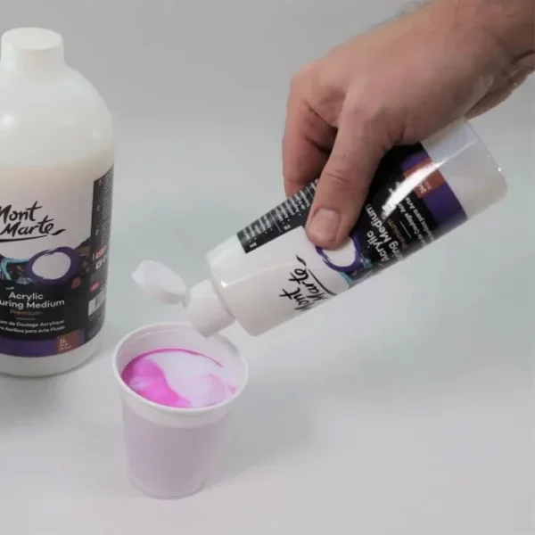 A persons hand is seen coming out of the right hand side of the frame, holding a bottle of Mont Marte Premium Pouring Medium. They are pouring it into a plastic cup that is filled with a bright pink paint. There is another bottle og Mont Marte Premium Pouring Medium coming in from the left hand side of the frame.