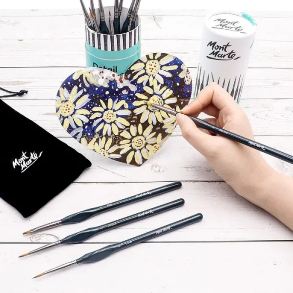 A persons hand is seen coming out of the right hand side of the frame, holding a brush from the Mont Marte Detail Brush Collection 15 piece. There are three brushes laying on the table in front of their hand. The brushes have black handles with the Mont Marte logo printed on them, and golden taklon bristles. They are painting a heart shaped canvas which is leaning up against the tube that the brushes come in. There is a black cloth bag coming out of the left hand side of the frame that is supplied in the tube set.