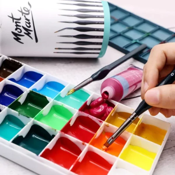 A persons hand is seen coming out of the right hand side of the frame, holding a paint brush from the Mont Marte Detail Brush Collection 15 piece. They are dipping the brush into some orange paint which is in a plastic palette. the palette is filled with a rainbow of different coloured paints. There is another brush laying on the palette and and open tube of pink paint against the palette.