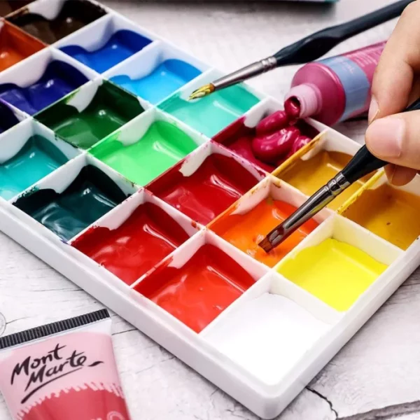 A persons hand is seen coming out of the right hand side of the frame, holding a paint brush from the Mont Marte Detail Brush Collection 15 piece. They are dipping the brush into some orange paint which is in a plastic palette. the palette is filled with a rainbow of different coloured paints. There is another brush laying on the palette and and open tube of pink paint against the palette.