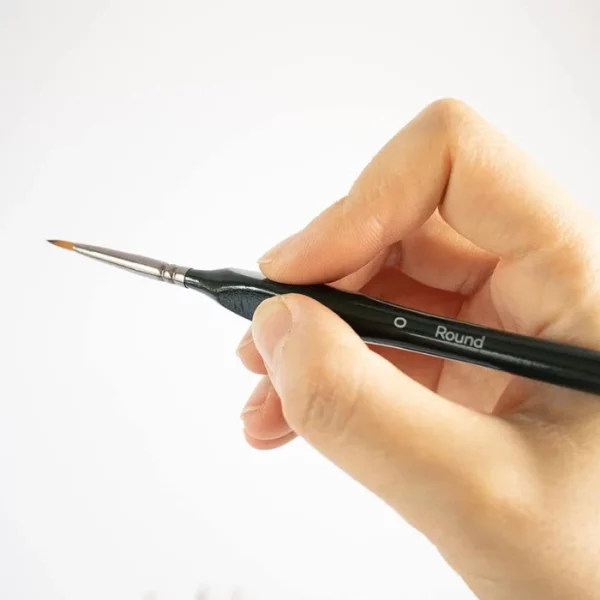 A persons hand is seen coming in from the right hand side of the frame. They are holding a single fine detail brush from the Mont Marte Fine Detail Brush Set. It is a close up of the hand and brush. The brush has a black handle and golden taklon bristles. On a white background.