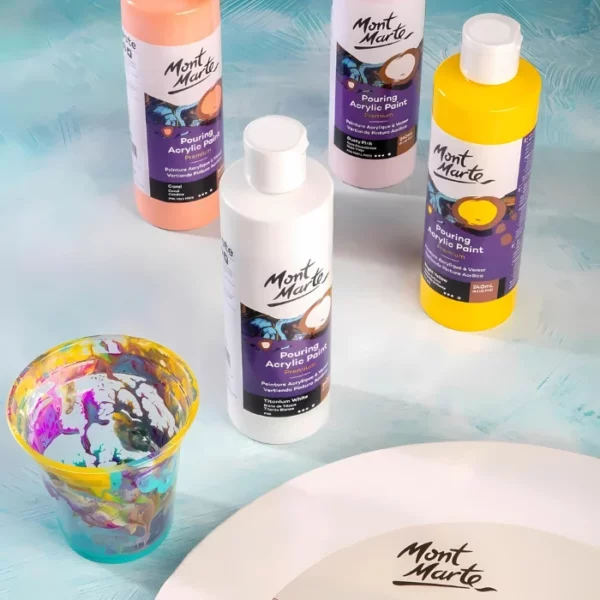 Four bottles of Mont Marte Pouring Acrylic Paint are standing on a desk with a used plastic cup that has had paint in it. Part of a Mont Marte round canvas can be seen coming in from the bottom of the frame.