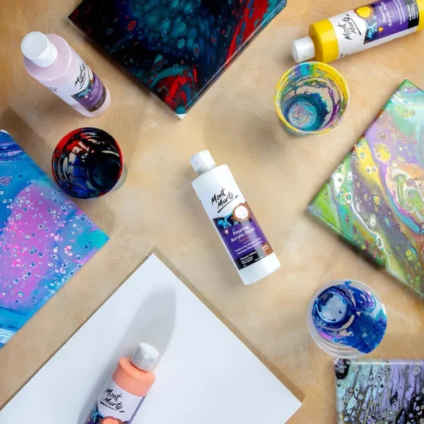 A birds eye view of a table with various bottles of Mont Marte Pouring Acrylic Paints and some painted canvases.