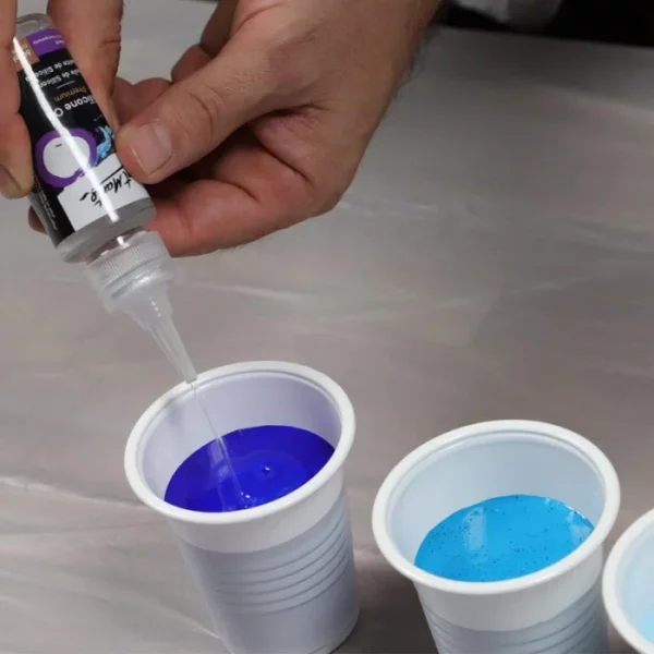 A persons hands are seen coming in at the top left hand corner of the frame. They are squeezing oil from a bottle of Mont Marte Premium Silicone Oil into a cup filled with blue paint. It is a plastic cup and there is another cup next to it, filled with a light turquoise paint.