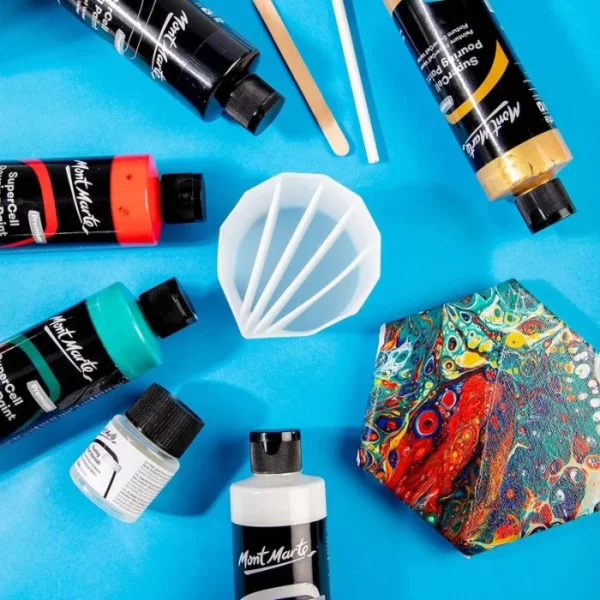 A Mont Marte Silicone Split Pouring Cup is shown sitting on top of a canvas that has been painted using the acrylic pouring technique. The cup is a white plastic in a rounded 'v' shape with four divisions to hold different coloured paints. There are numerous small bottles of paint scattered around the canvas.