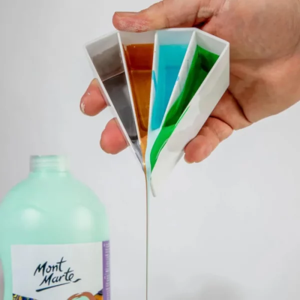 A persons hand is seen coming out of the top right hand corner of the frame, holding a Mont Marte Split Pouring Cup with paint in it, and they are pouring it. There are 4 colours of paint in the white plastic cup. You can see a tall bottle of turquoise paint in the bottom left hand corner of the frame.