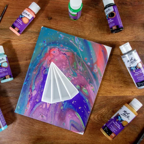 A Mont Marte Split Pouring Cup is shown sitting on top of a canvas that has been painted using the acrylic pouring technique. The cup is a white plastic in a 'v' shape with four divisions to hold different coloured paints. There are numerous small bottles of paint scattered around the canvas.