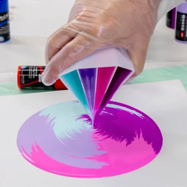 A persons hand is seen coming out of the top of the frame, holding a Mont Marte Split Pouring Cup with paint in it, and they are pouring it. over a white canvas and the pour is forming a circle of paint on the canvas. There are 4 colours of paint in the white plastic cup.