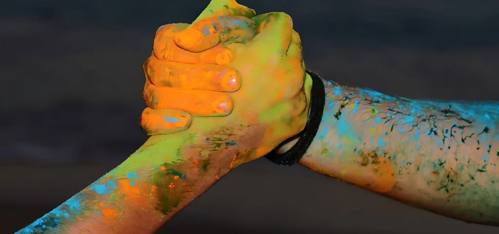 two peoples hands are visable giving eachother a paint covered handshake.. their hands are covered in shades of orange, yellow and blue and the person on the right hand side has a bracelet that is black on. the background is black and it is in the center of the image.