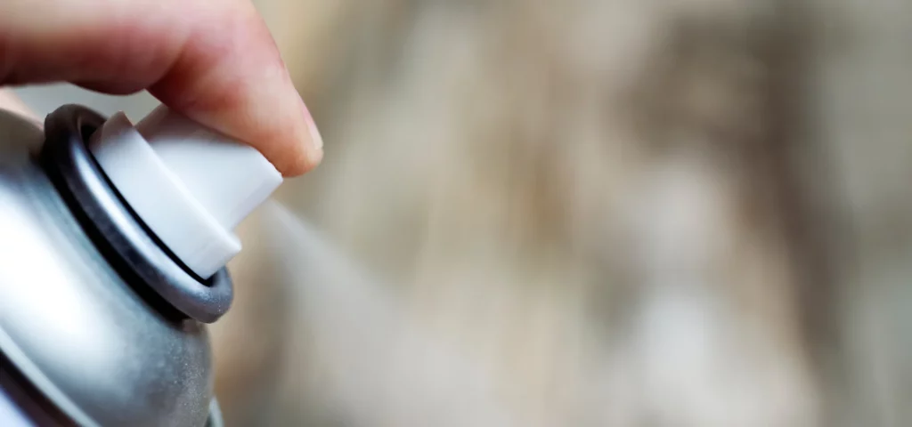 the image is of spray adhesive in action. there is a close up of a persons finger holding onto the nozzle of the spray can which is white and pressing down onto it which is making the adhesive come out of it. the can has a white nozzle, with a silver and white body of the can. the background is blurred but is of the outside area