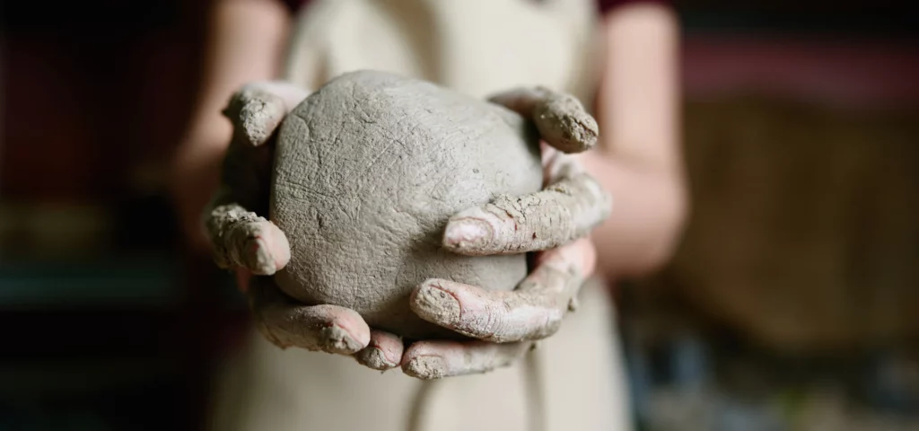 in the center of the image you can see a woman s hands covered in clay holding a clay ball that is a grey colour. they clay ball is wet and cracked and is being used. her hands are full of clay and in the background in a blur you can see her torso in an apron that is a beige colour.